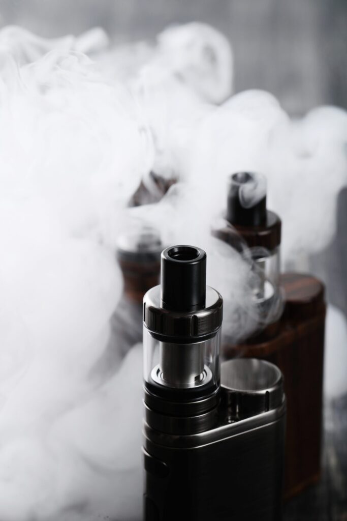 Close-up of a vaping device with an atomizer on top, surrounded by a dense cloud of vapor, against a blurred background of similar devices.