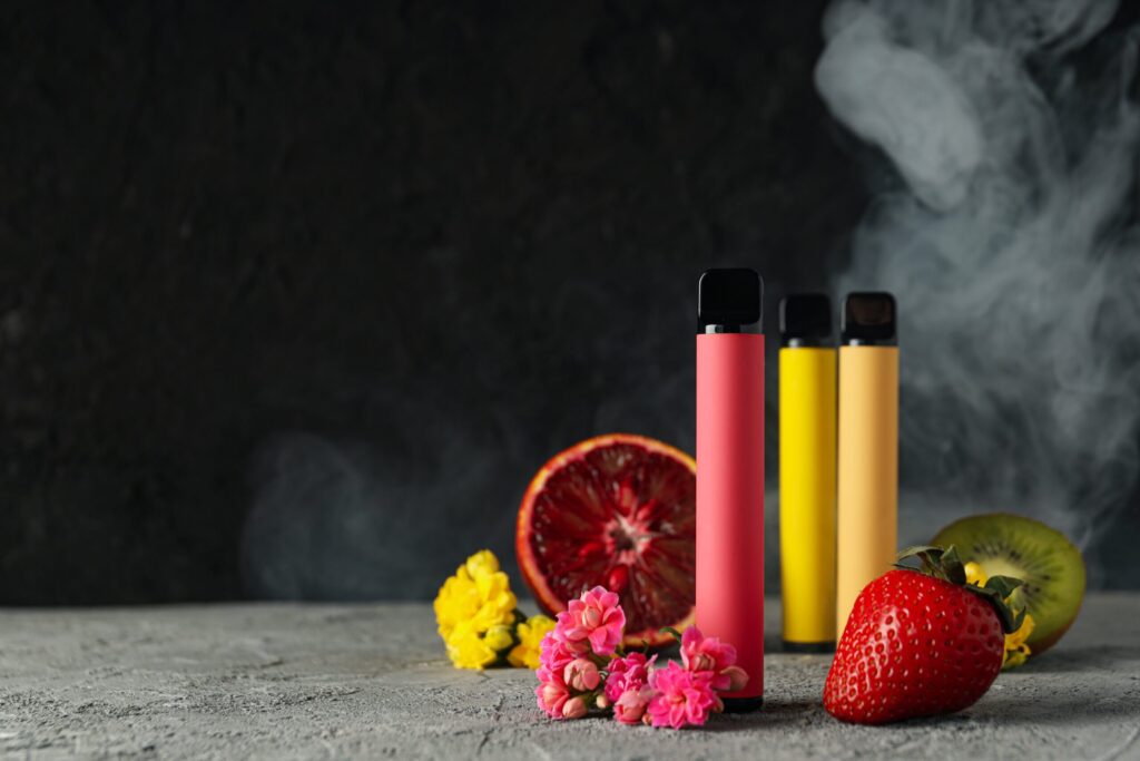 Three colorful vaping devices stand upright next to a halved blood orange, fresh strawberries, and vibrant flowers, with a wisp of vapor in the background.