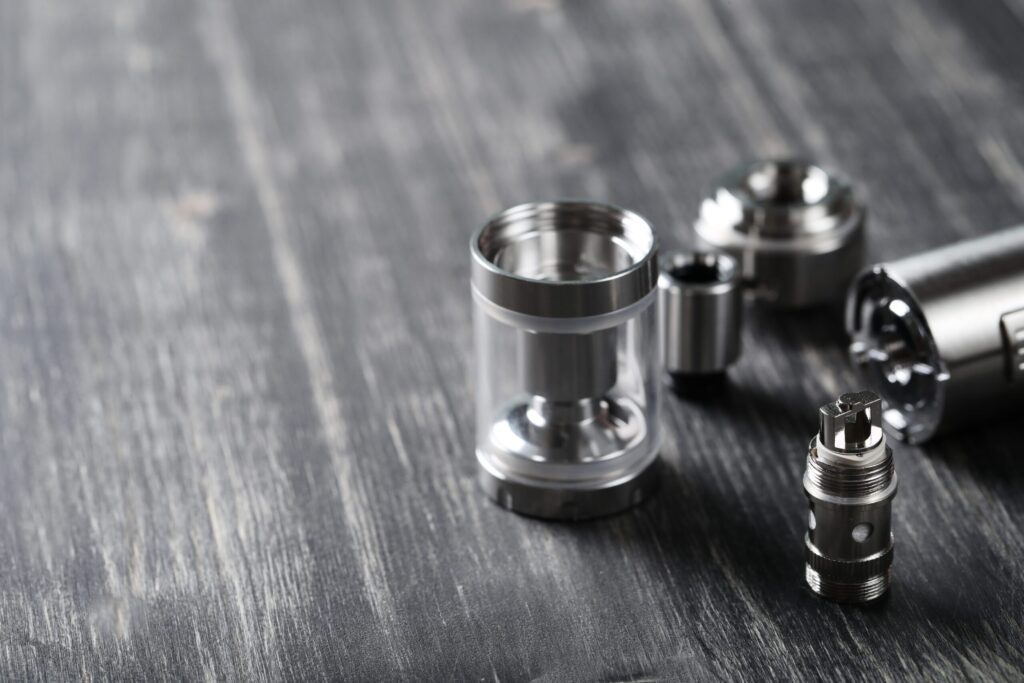 Several metallic vaping tank components are arranged on a dark wooden surface, showcasing different parts of a vape setup.