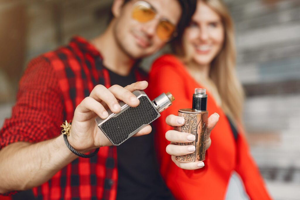 A young couple holding up two vaping devices to the camera, the man with sunglasses and a checkered shirt, and the woman in a red top smiling in the background.