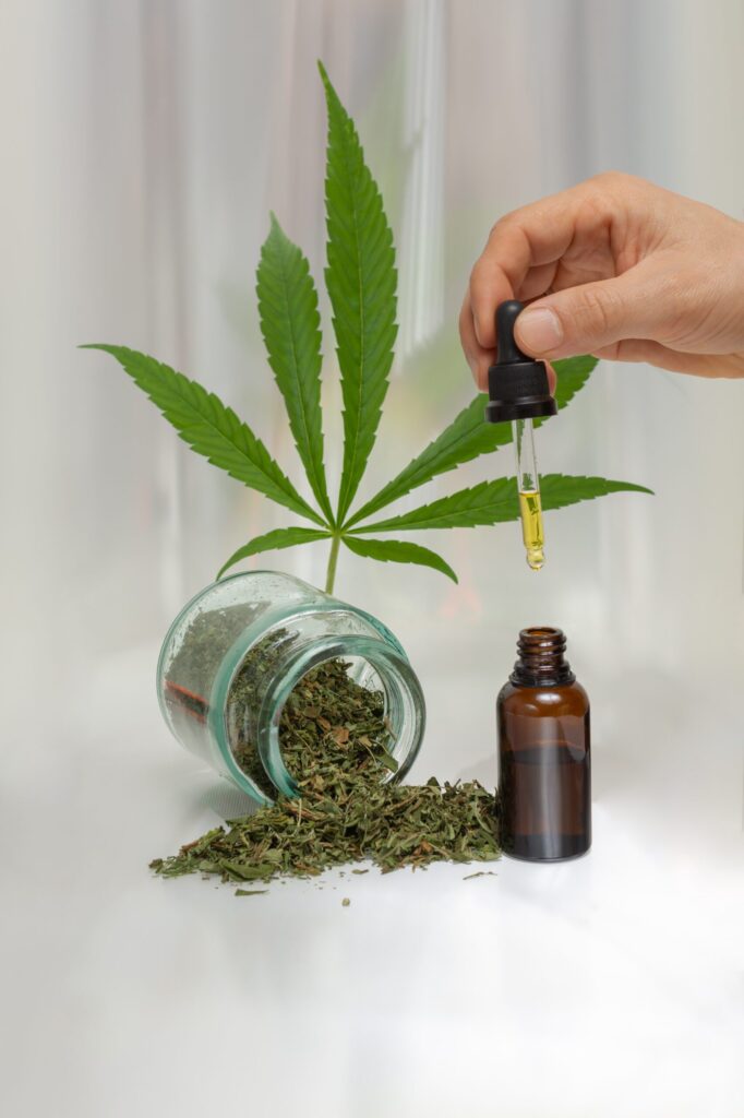 A hand using a dropper to dispense CBD oil with a cannabis leaf and dried herbs in the background.