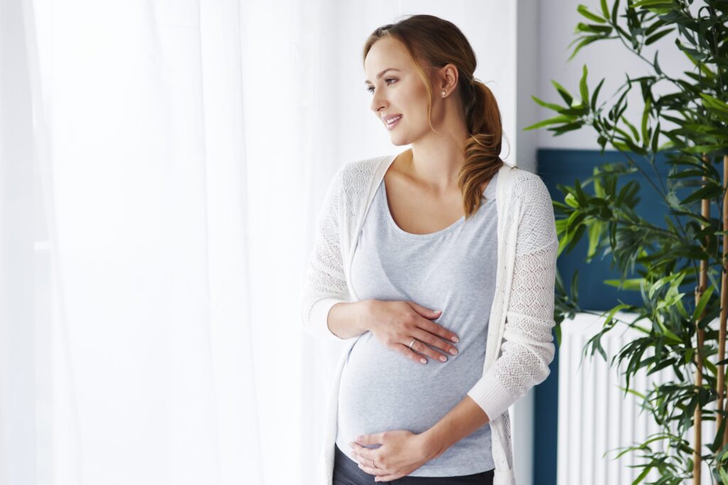 A pregnant woman gently cradling her belly while looking out a window.